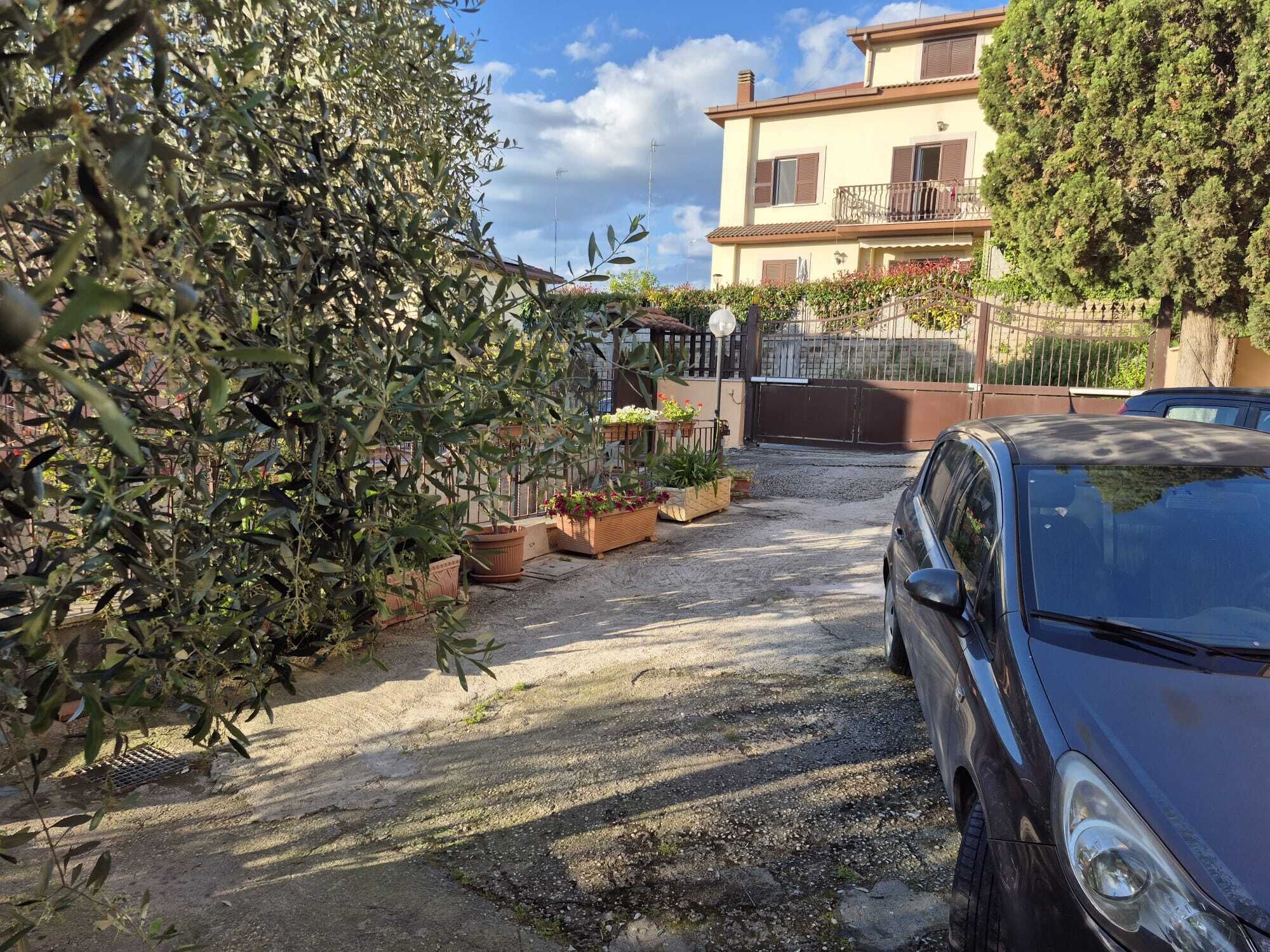 A paved driveway leading to a building with a gated entrance, surrounded by potted plants and a parked car.