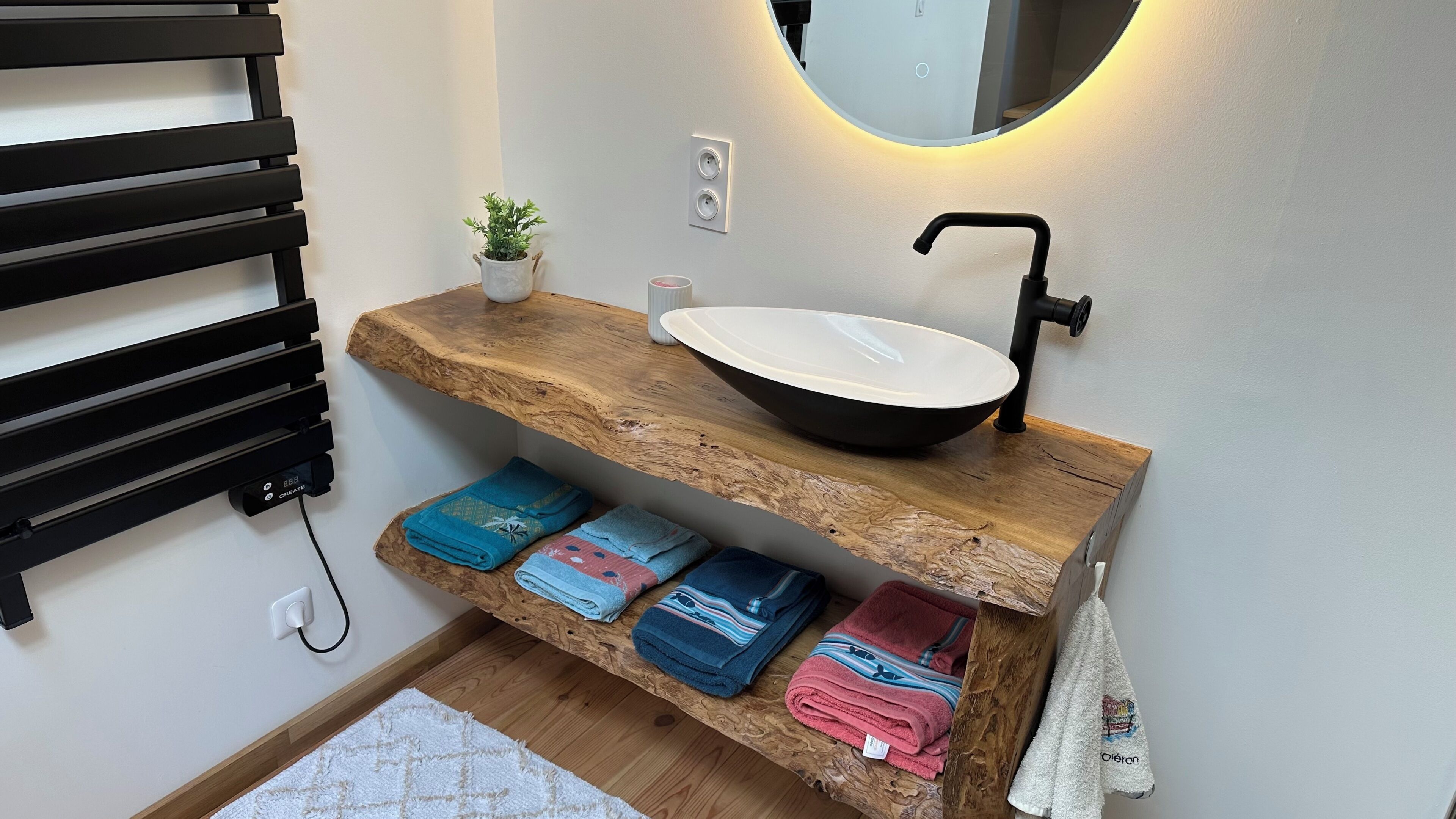 Une salle de bain avec un meuble-lavabo en bois, un miroir rond, un robinet noir et une étagère où sont pliés des serviettes.