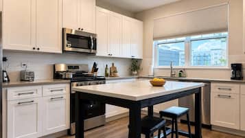 A modern kitchen with white cabinetry, a central island with stools, a microwave, a stove, and a window with blinds.
