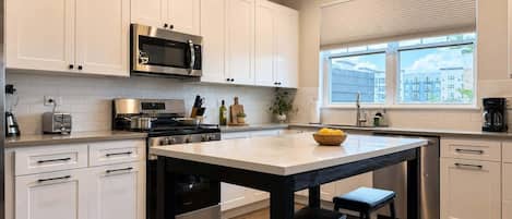 A modern kitchen with white cabinetry, a central island with stools, a microwave, a stove, and a window with blinds.