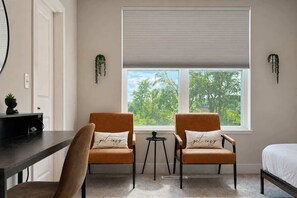 Une pièce comprenant deux fauteuils, une petite table, un lit et une fenêtre avec des stores.
