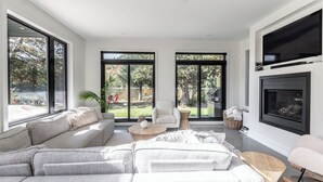 Uma sala de estar moderna com um sofá modular cinza, uma poltrona branca, uma mesa de centro de madeira e uma lareira.