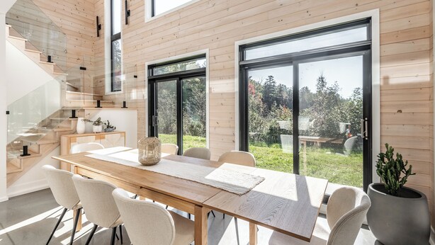 Um interior moderno com piso de madeira, uma mesa de jantar com cadeiras, uma escada e janelas amplas.