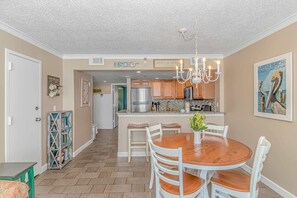 A dining area with a wooden table and white chairs, a kitchen in the background, and a hallway leading to another room. - Sand Dunes 1043 (Myrtle Beach)