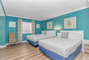 A room with two beds, a wicker chair, a window with curtains, and wall art. - Sand Dunes 1043 (Myrtle Beach)