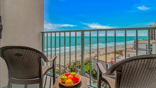Un balcon avec des fauteuils en osier, une table sur laquelle est posé un panier de fruits, et une vue sur la plage et l’océan.