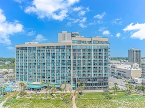A multi-story hotel with a modern glass facade, a swimming pool, and a landscaped courtyard. - Sand Dunes 1044 (Myrtle Beach)