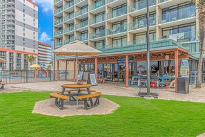 Un restaurante frente a la playa con terraza al aire libre, un porche cubierto y vista a un edificio alto al fondo.