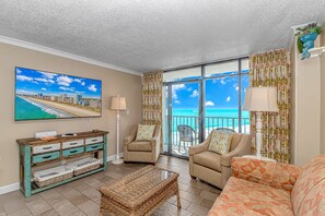A living room with a large TV, a sofa, a coffee table, and a view of the ocean through large windows. - Sand Dunes 1043 & 1044 (Myrtle Beach)