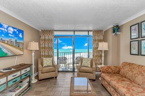 A living room with a sofa, two armchairs, a coffee table, a TV, and a balcony with a view of the beach and ocean. - Sand Dunes 1043 & 1044 (Myrtle Beach)