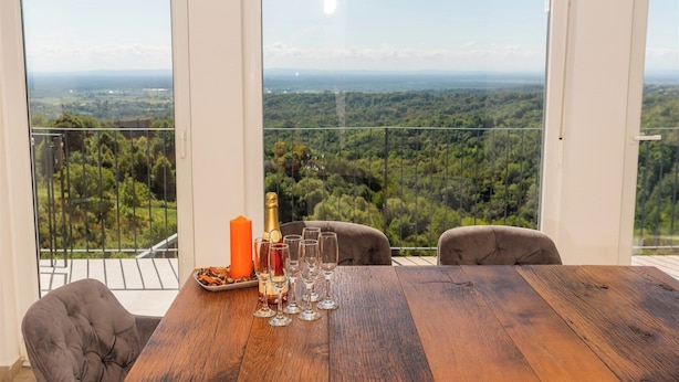 Un balcón con una mesa preparada para una comida, una botella y copas, con vistas a un paisaje boscoso.