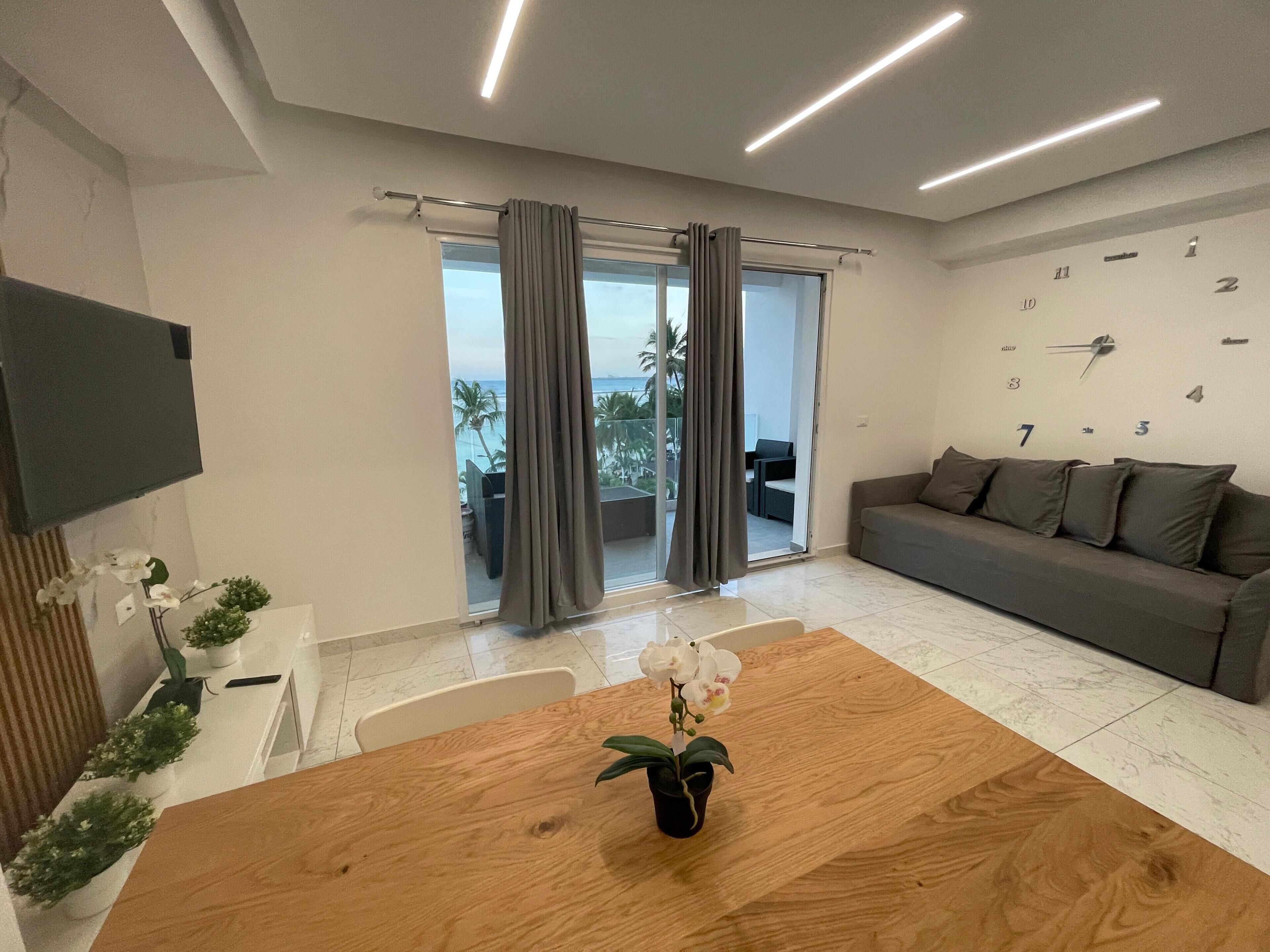 Una sala de estar moderna con una mesa de comedor de madera, un sofá gris y un ventanal con cortinas.