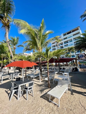 Een strandgebied met ligbedden en parasols, palmbomen en een gebouw met een bord waarop 'PIZZERIA' staat.