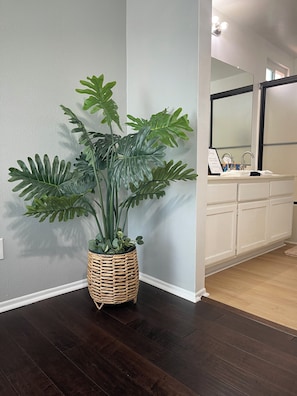 Un rincón con una planta grande en maceta, un baño blanco con espejo y lavabo, y suelo de madera oscura.