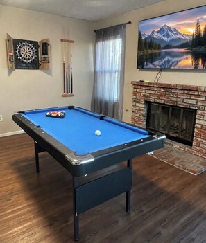 A pool table with a single white ball, a dartboard, and a fireplace with a mountain landscape painting above it.