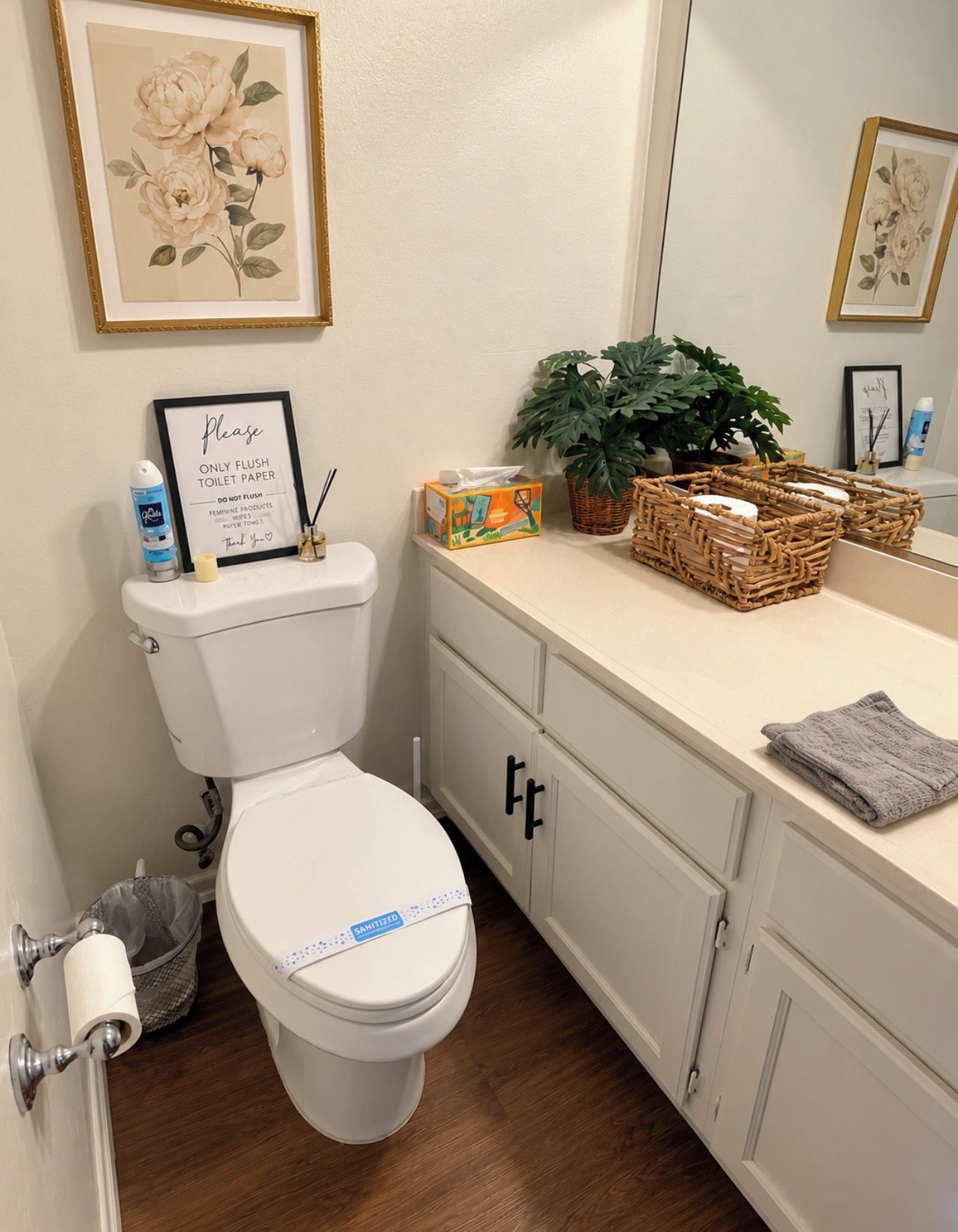 A bathroom with a toilet, a sink with a mirror, a framed sign, a plant, and a basket.