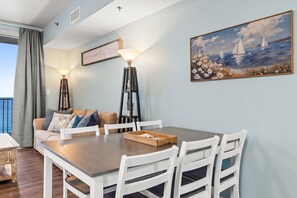 A dining area with a wooden table and white chairs, a sofa with cushions, a painting of sailboats, and a sign on the wall. - Shores of Panama - Gulf Views - 2BR + Bunks - Beach Chairs Included (Panama City Beach)