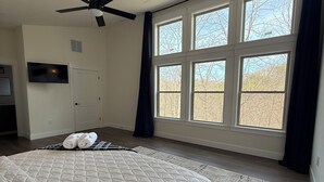 A bedroom with a large bed, a ceiling fan, a TV, and a view of trees through large windows. - RRG Cabin - 2 King Suites + Ride-In Access (Stanton)