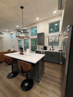 A modern kitchen with a central island, wooden chairs, a hanging light fixture, and a large window.