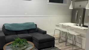 A modern living room with a sectional sofa, a round coffee table with a plant, and a kitchen area with a white countertop and stainless steel appliances. - Cozy two-bedroom basement apartment equipped with a full kitchen! (Min 28 days) (Lake Loon)