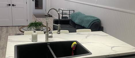 A kitchen with a sink, cabinets, a dishwasher, pendant lights, a potted plant, and a teal throw blanket on a sofa.