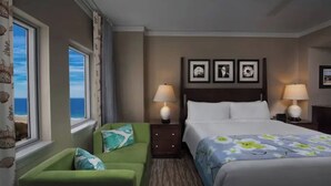 Habitación de hotel con cama, dos mesitas de noche con lámparas, un sillón verde y vista al mar.