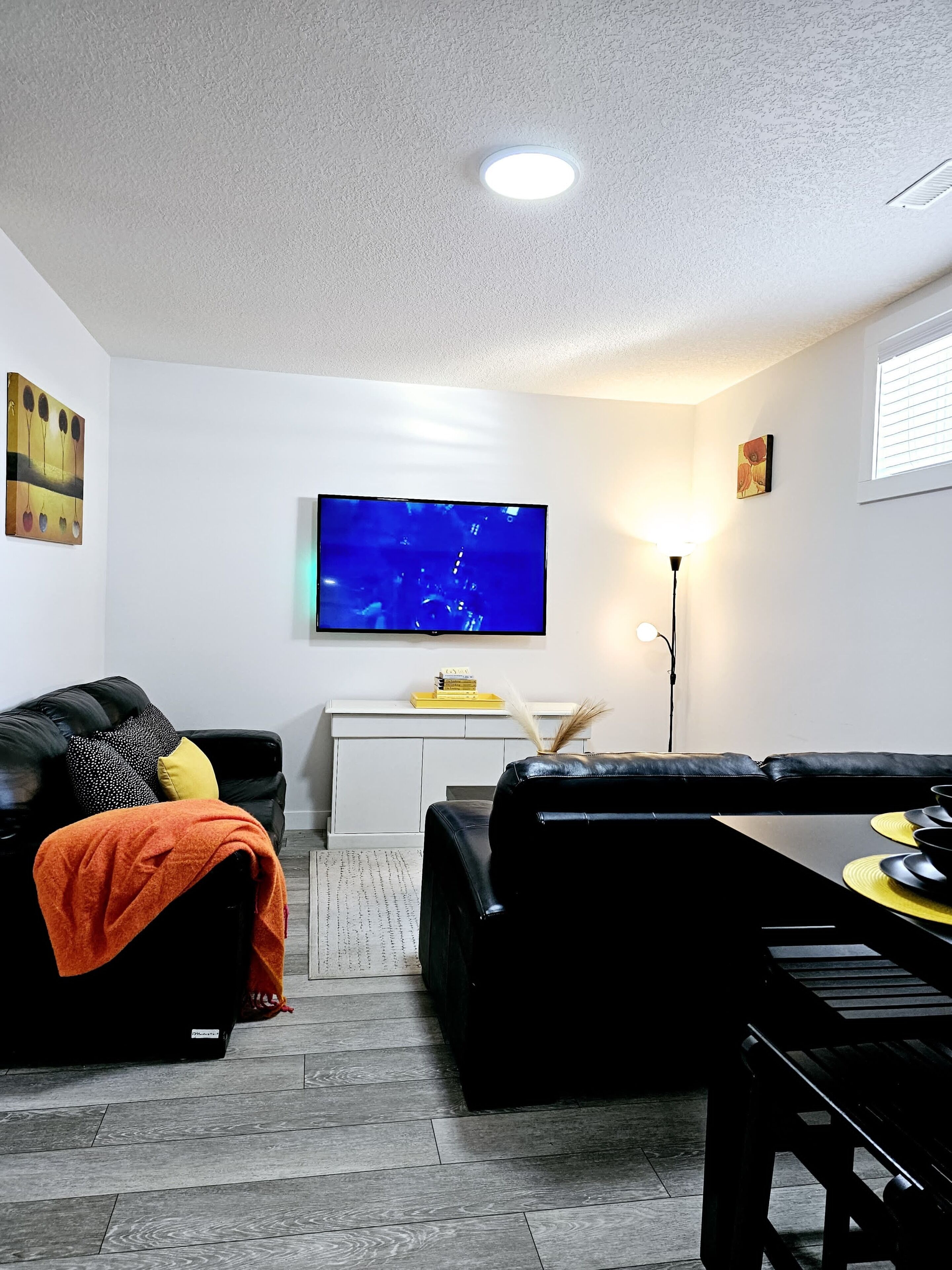 Ein Wohnzimmer mit einer schwarzen Ledersofa, einem Esstisch mit gelben Platzsets, einem an der Wand montierten Flachbildfernseher und einer weißen Kommode darunter.