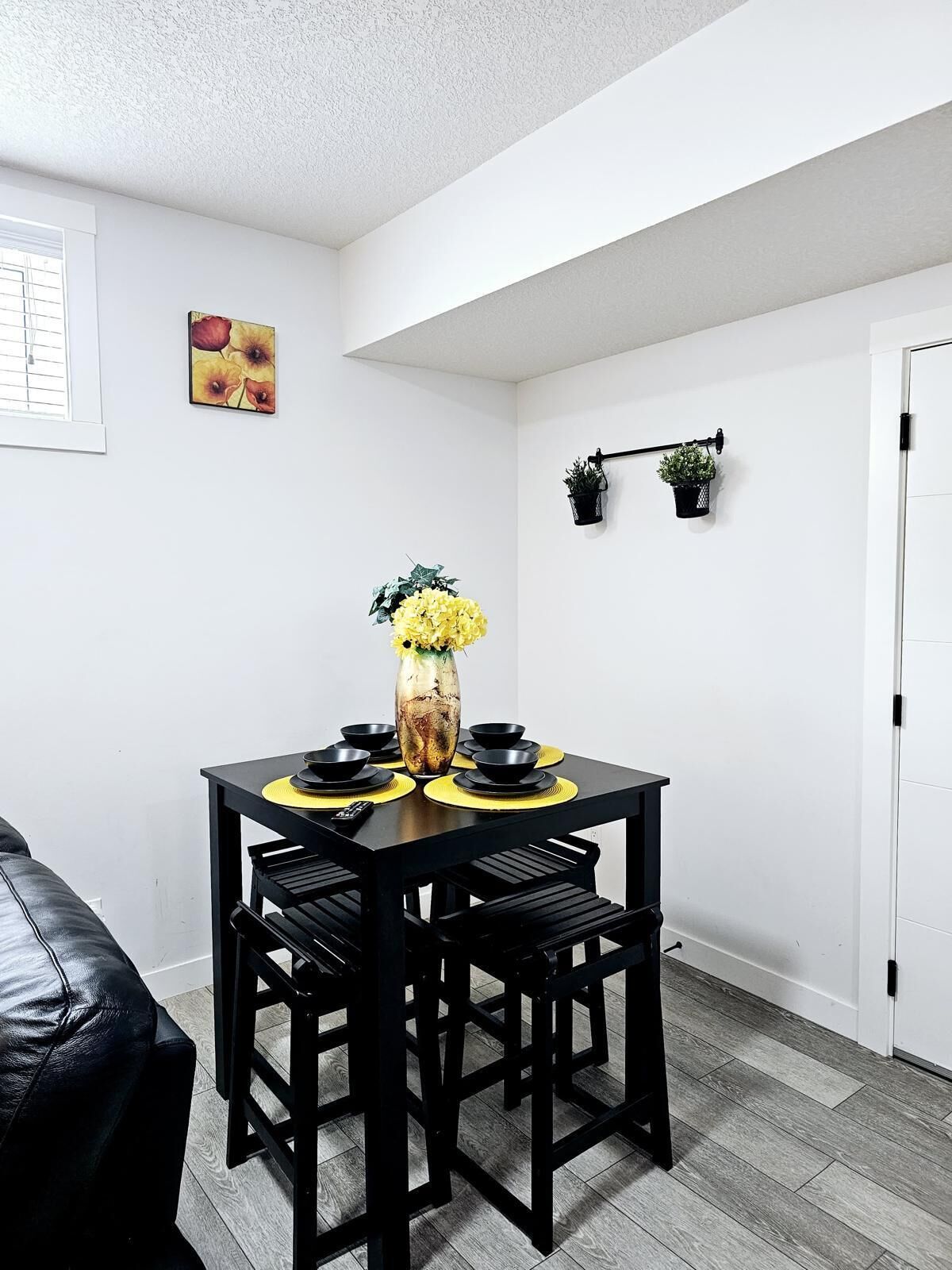 Un comedor con una mesa y sillas negras, un florero con flores amarillas y dos plantas en macetas en la pared.