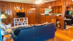 Una sala de estar con un sofá azul, paredes de madera, un televisor y una zona de cocina.