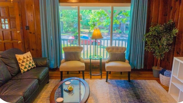 Una habitación con puerta de madera, un sofá de cuero negro, dos sillones, una mesa de centro de vidrio, una lámpara y vistas a un jardín a través de amplios ventanales.