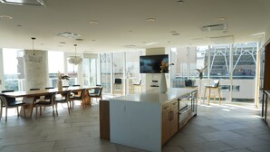A modern kitchen with a central island, a dining area with chairs, a TV mounted on the wall, and large windows offering a view of the city.