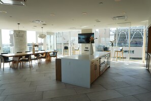 Une cuisine moderne dotée d’un îlot central, d’un coin repas avec des chaises, d’un téléviseur fixé au mur et de grandes fenêtres offrant une vue sur la ville.