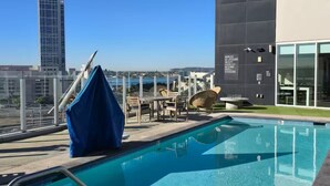 A rooftop pool with a view of a city skyline, a covered seating area, and a building with a lifeguard warning sign.