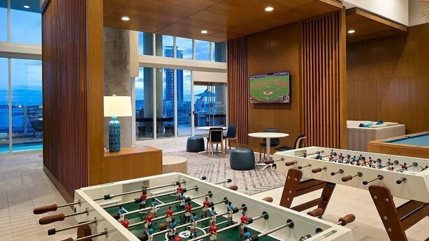 A modern indoor space with two foosball tables, a pool table, and a large window offering a view of the cityscape.