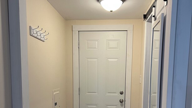 A hallway with a white door, a light fixture on the ceiling, a coat rack, and a wooden floor.