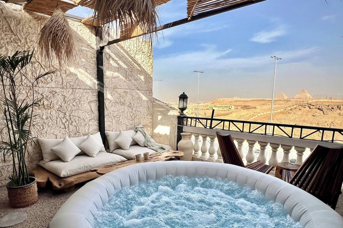 A room with a hot tub, a sofa with cushions, a potted plant, and a view of pyramids.