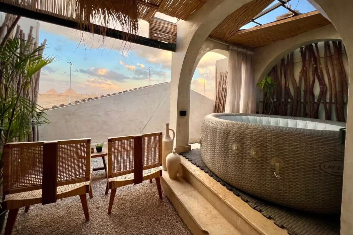 A room with a hot tub, two wicker chairs, a view of pyramids, and a thatched ceiling.