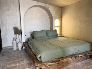 Un dormitorio con estructura de cama de madera, colcha verde y dos jarrones con plantas secas.