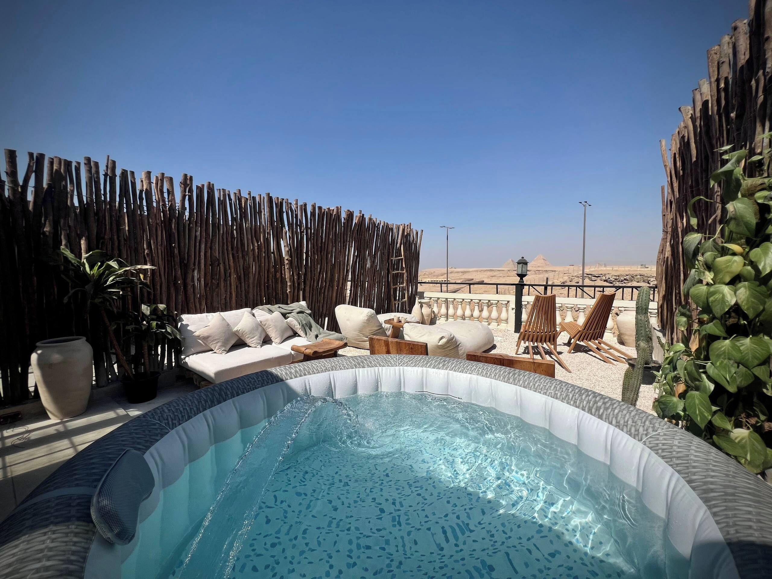 A hot tub surrounded by outdoor furniture, a wooden fence, and a view of pyramids.
