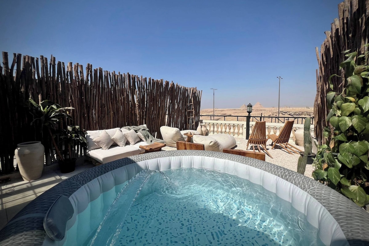 A hot tub surrounded by outdoor furniture, a wooden fence, and a view of pyramids.