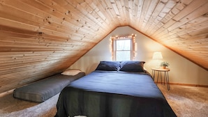 Tunnelmallinen ullakkohuone, jossa on kalteva katto, tummilla tekstiileillä varustettu sänky, yöpöytä lampun kanssa ja pieni penkki.
