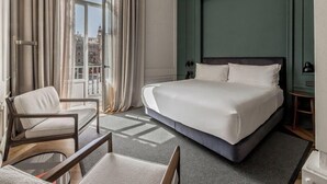 A hotel room with a large bed, a chair, a small table, and a view of the city through the window. - Mercer Plaza Sevilla (Seville)