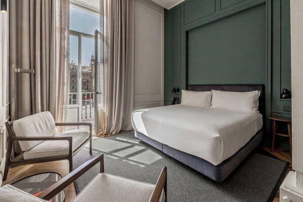 A hotel room with a large bed, a chair, a small table, and a view of the city through the window. - Mercer Plaza Sevilla (Seville)