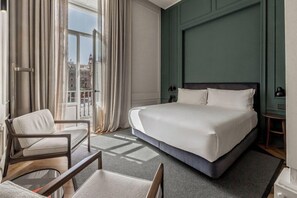 Hotellihuone, jossa on suuri sänky, tuoli, pieni pöytä ja ikkunasta avautuva näkymä rakennuksiin.