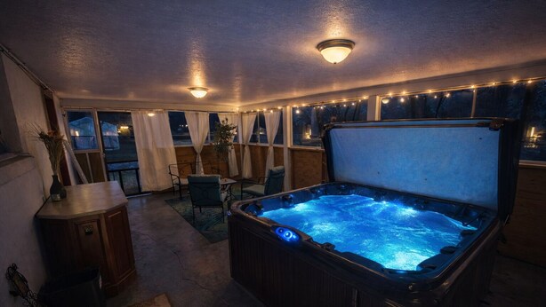 A hot tub with a wooden exterior and a blue light, surrounded by windows with curtains, a small table, and chairs. - Peaceful Hot Tub Hideaway - 15 mins to Chatt - Dog Friendly (Rossville)
