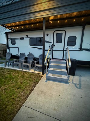 A covered outdoor area with a concrete staircase leading to a white RV with gray chairs and a table. - Enjoy a cozy getaway in this modern 1-bedroom place. (Rosenberg)