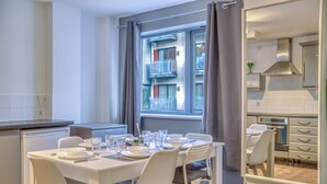 A modern kitchen with white cabinets, a dining table set for four, and a large window with curtains. - The Manchester City Haven (Manchester)