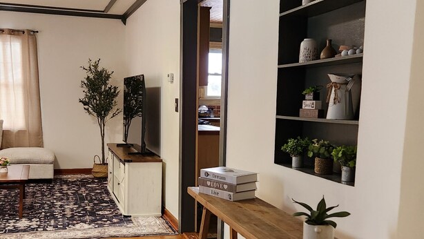 A room with a wooden floor, a checkered rug, a wooden console table, a shelf with decorative items, and a doorway leading to another room. - ​Upstate Oasis: Moody Chic Interior & Summer Pool Fun (Carthage)