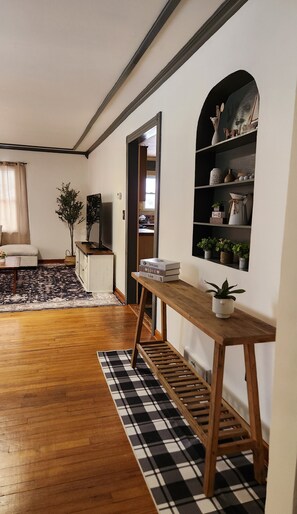 A room with a wooden floor, a checkered rug, a wooden console table, a shelf with decorative items, and a doorway leading to another room.
