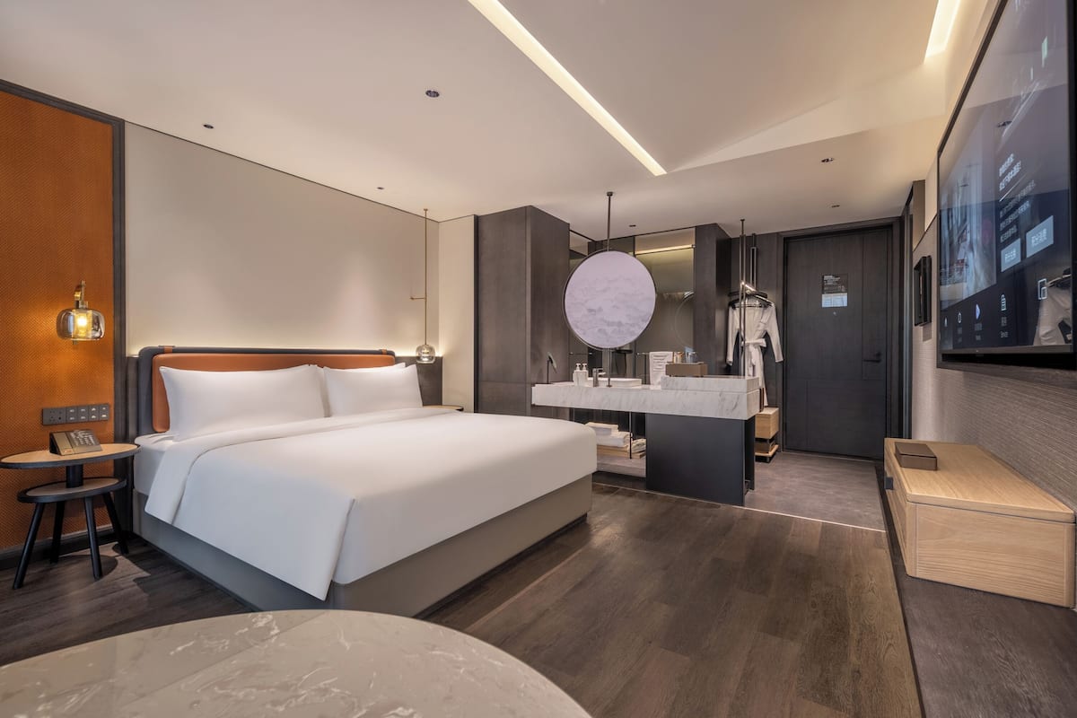 Une chambre d’hôtel moderne avec un grand lit, une petite table de chevet, un miroir rond, une coiffeuse intégrée avec un miroir et une commode en bois.